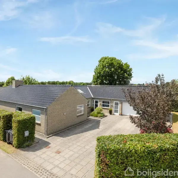 Et moderne, fladt-tag hus står stille omgivet af hække. Det er placeret ved siden af en stille vej med en flisebelagt indkørsel og grønne områder i baggrunden.