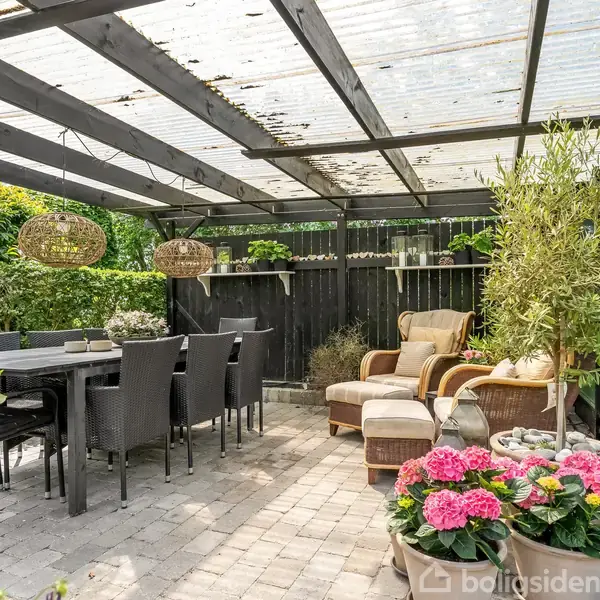 Terrasse med spisebord og stole under glasoverdækning. Ved siden af står en loungestol med skammel. Blomster og planter omkranser området, mens flettede lamper hænger fra tagkonstruktionen.