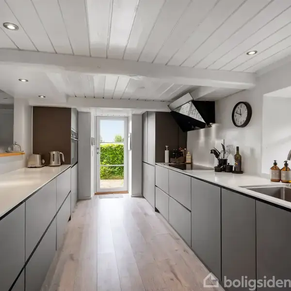 Køkken med grå skabsfacader og hvidt loft, i en stilfuld moderne indretning. Rummet er lyst med vinduer over vasken og en åben dør mod en grøn have i baggrunden.