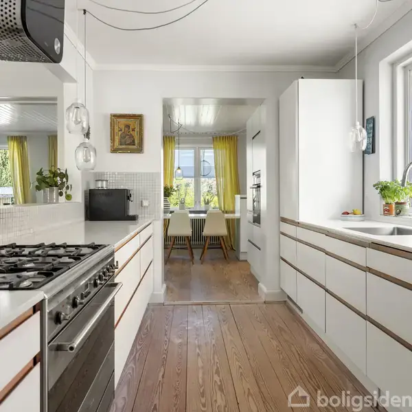 Komfur står i et lyst køkken, omgivet af hvide skabe, med en spiseplads i baggrunden, der har lyse gardiner og et bord ved vinduet.
