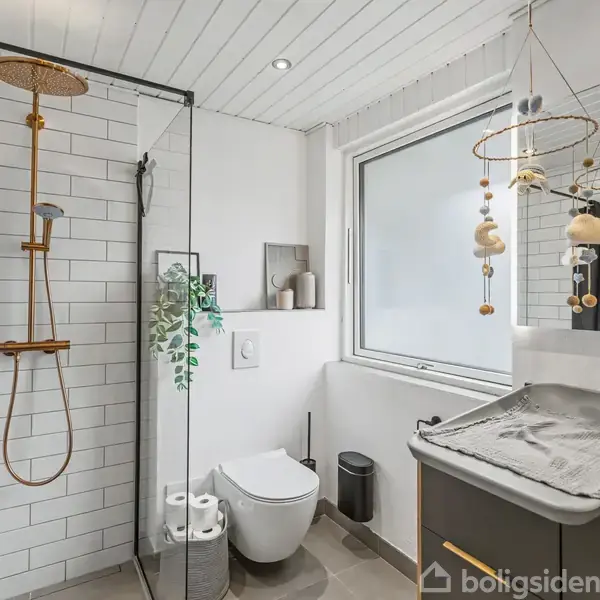 Bruser i glasinddelt kabine med hvide fliser og kobberarmatur, toilet ved siden af. Vægophængt puslebord under et vindue, arrangeret med barnelegetøj over det.