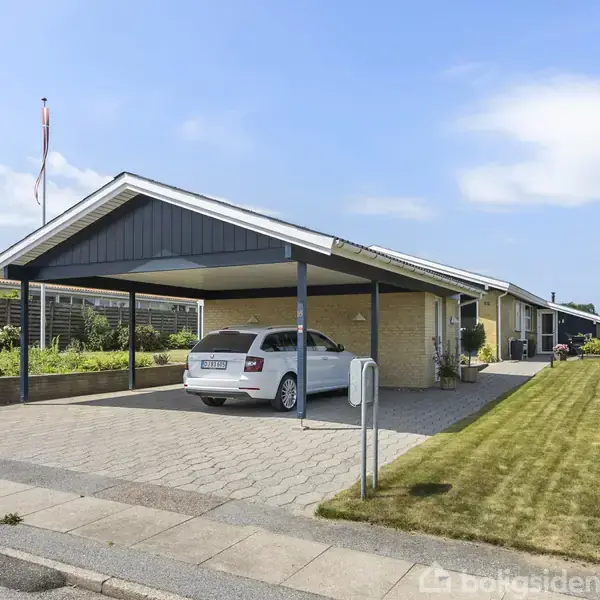 Carporten har en hvid bil parkeret, og den ligger ved siden af en moderne, gulstensbungalow. Omgivet af velplejet græsplæne og hæk i et stille boligområde.