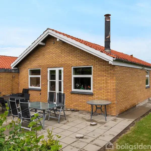 En gul murstenshus står på en flisebelagt terrasse med havemøbler i en have. Huset har rødt tegltag og flere vinduer samt en skorsten.