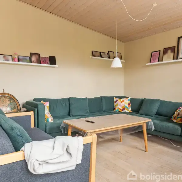 En grøn hjørnesofa står i en stue med lamper over et træbord. Væghylder med billeder pryder væggene, og en jordklode står ved siden af sofaen.