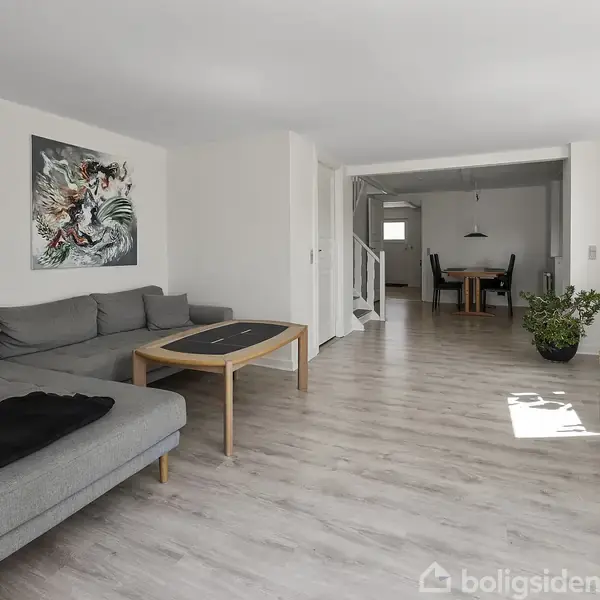Grå sofa står stille i en moderne stue med lyse trægulve, kunst på væggen og et stort vindue til højre. I baggrunden ses et spiseområde med stole.