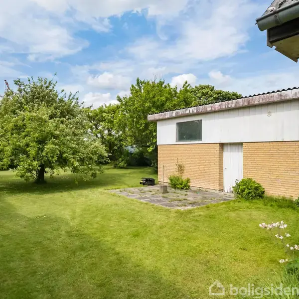 En gulstensbygning med en flad tag fremstår i en grøn have, hvor et stort træ skaber skygge. Haven er godt vedligeholdt med græsplæne og buske.