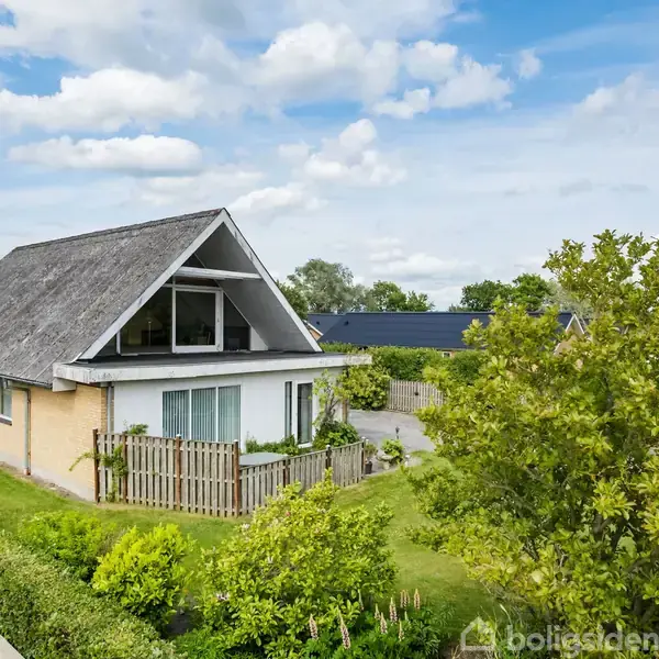 Hus med stråtækt tag står roligt omgivet af grønne buske og træer. En lille have er foran huset, og en brostensbelagt vej løber på den ene side under en skyfri himmel.