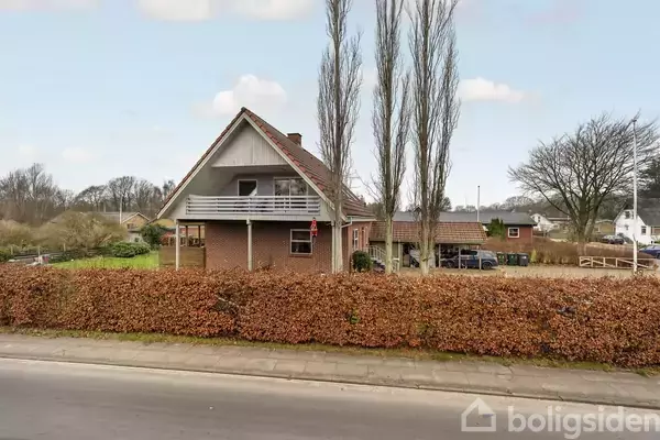 Et rødt murstenshus har en bred balkon og omgives af en hæk. To høje træer står foran huset, som ligger i et villakvarter med flere huse og træer i baggrunden.