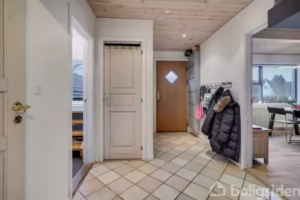 Gang med fliser, hvor jakker hænger på kroge til højre. En lukket trædør er forenden, omgivet af lyse vægge og loft. Døre fører ind til andre rum.