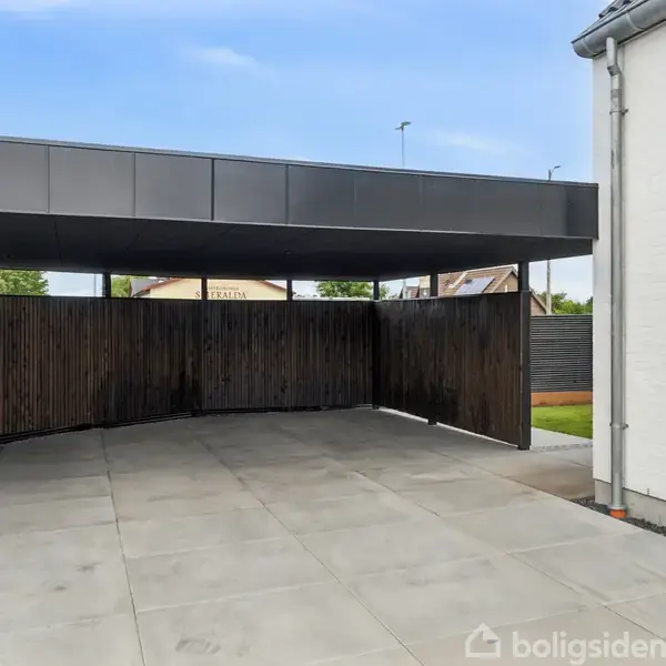 Carport med fladt, moderne tag står ubrugt. Fliser danner gulv, og mørke træplanker udgør bagvæggen. Til højre ses en hvid husmur. Himmel er klar og blå.