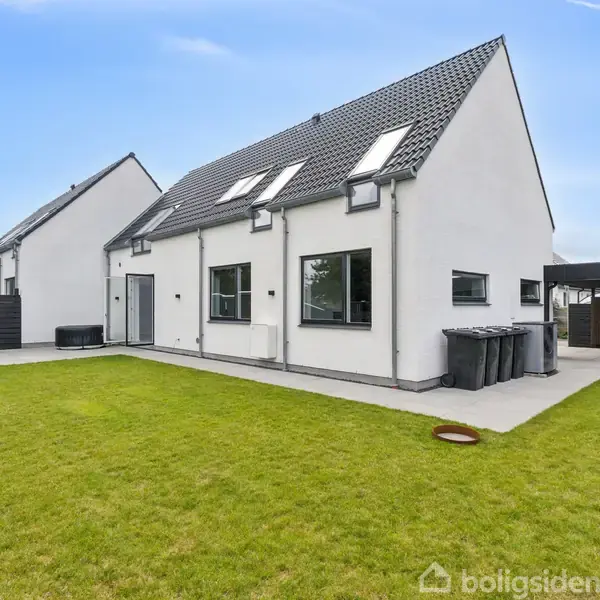 En moderne, hvid villa med skråt tag står stille i en velplejet have. Villaen har store vinduer og er omgivet af en betonsti og en carport i nærheden.