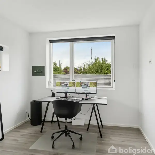 Et skrivebord med to skærme står ved et vindue i et lyst hjemmekontor. En kontorstol er placeret foran bordet, og en stor plakat står til venstre.