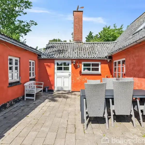 Rødt murstenshus omkranser en brostensterrasse med et stort sort havebord og seks stole. Dør og vinduer er hvide. Træer ses i baggrunden under en klar blå himmel.