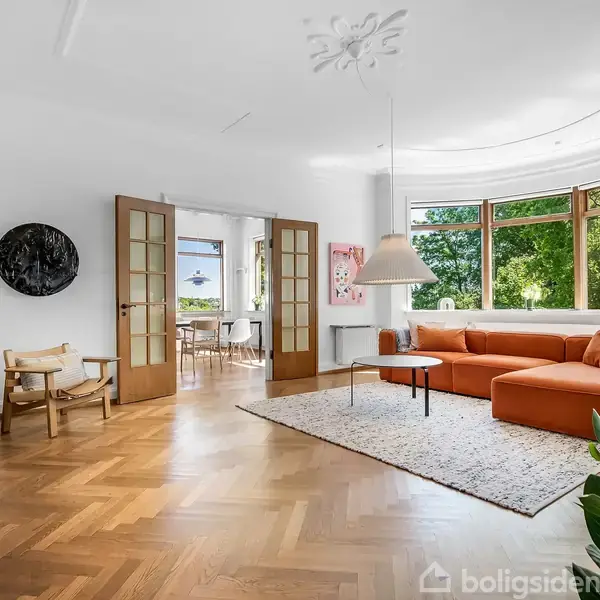 En stue med en orange sofa står på et tæppe. Rummet har buede, store vinduer mod en grøn have. Der er også træmøbler og planter, og et spiseområde ses i baggrunden.