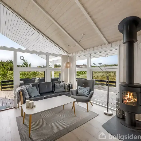 Brændeovn i hjørnet af en lys stue, hvor den er tændt og afgiver varme. Ved siden af står en moderne sofa og en stol. Store vinduer giver udsigt til en terrasse og grønne omgivelser.