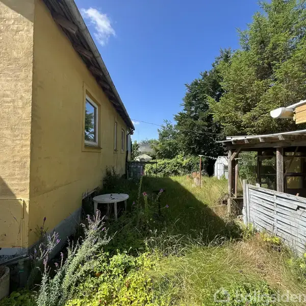 En gul bygning står stille i solen ved siden af et græsareal med høje urter og en lille stibord. Til højre er en overdækket terrasse og høje træer i baggrunden.
