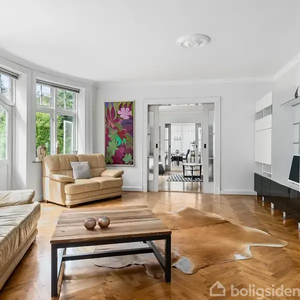 Stue med beige sofaer og sofabord, dekoreret med kunst på væggen. Stor vinduesvæg slipper naturligt lys ind. Til højre ses en moderne reol. I baggrunden ses en spisestue.