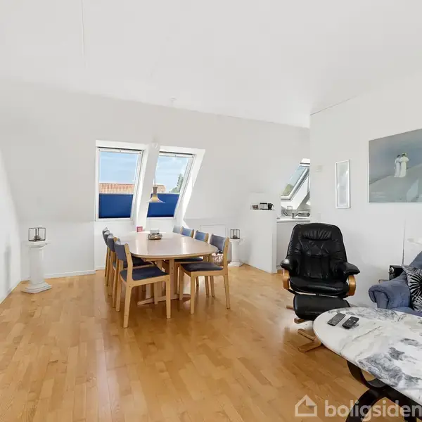 En stue med trægulv, hvor et spisebord med stole står foran to store skråvinduer. I højre side er en sort lænestol ved siden af en grå sofa og et sofabord.