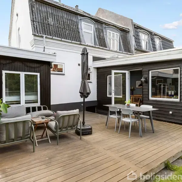 Træterrasse med møbler og parasol står foran et sort træhus. Et hvidt hus med kvistvinduer ses i baggrunden, og der er grønne planter til venstre.