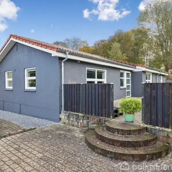 Grå villa med rødt tag står stille i et lukket haveområde med stentrappe. Hvide skodder omkranser de store vinduer. Baggrunden består af træer og blå himmel.