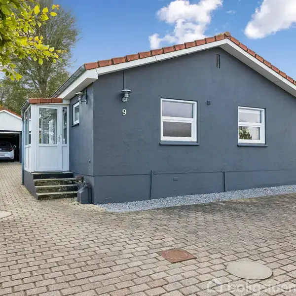 Et gråt hus med rødt tag og nummer 9 på facaden står ved en brolagt indkørsel. Til venstre er der en garage med en bil indeni, omgivet af grønne buske.