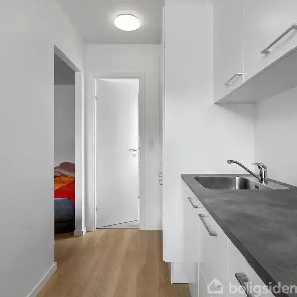 Et hvidt køkken med en enkelt vask og en grå bordplade. I baggrunden ses en dør til et værelse med en seng.