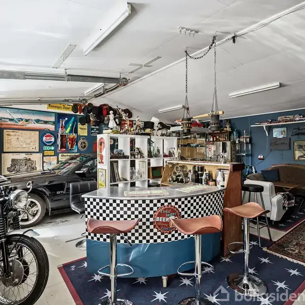 Garage indrettet som en retro bar med et rundt barområde i checkerboard-design og tre barstole. En motorcykel står ved siden af, omgivet af biltilbehør og vintage plakater.