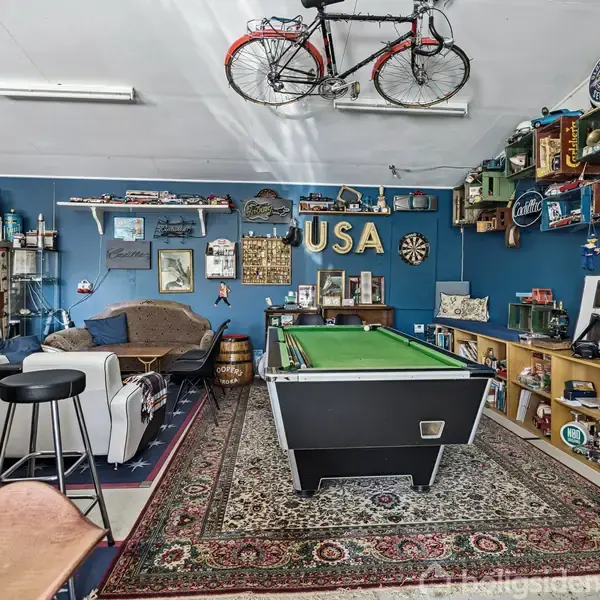 En stue med en poolbord i centrum under en hængende cykel, omgivet af sofaer, barstole og hylder fyldt med samlerobjekter. "USA" står skrevet på den blå væg.