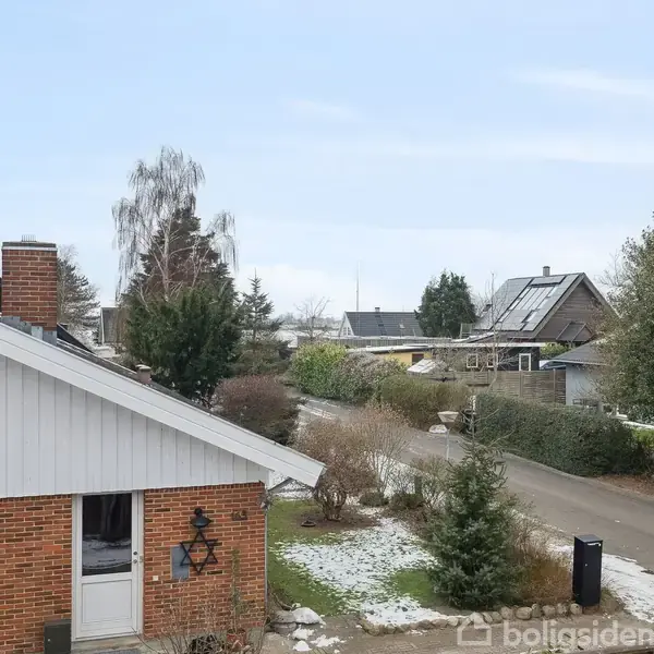 Et hus ligger ved en stille vej omgivet af andre huse og træer. Let sne dækker nogle områder af jorden, mens himlen er lys og klar.