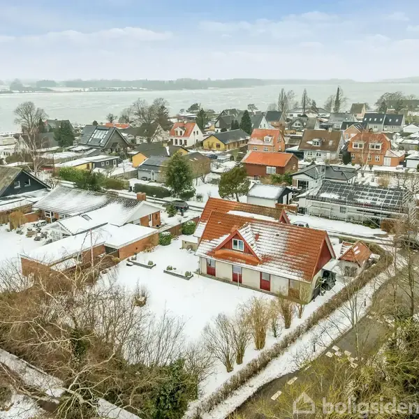 Røde hustage er dækket af sne i en bebyggelse ved vandet. Sneklædte haver og veje omkranser husene under en overskyet himmel. Ingen synlig tekst.