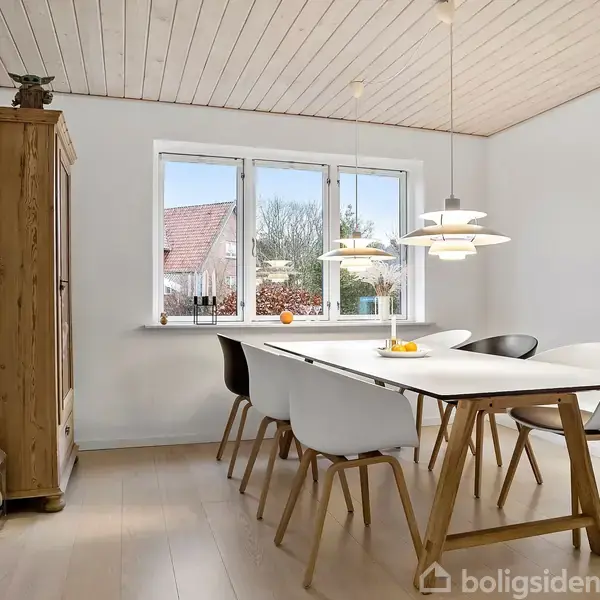 Et spisebord med seks stole står i et lyst moderne rum med træloft. Der hænger lamper over bordet, og et vindue viser et udsyn til huse og træer udenfor. En træskab står til venstre.