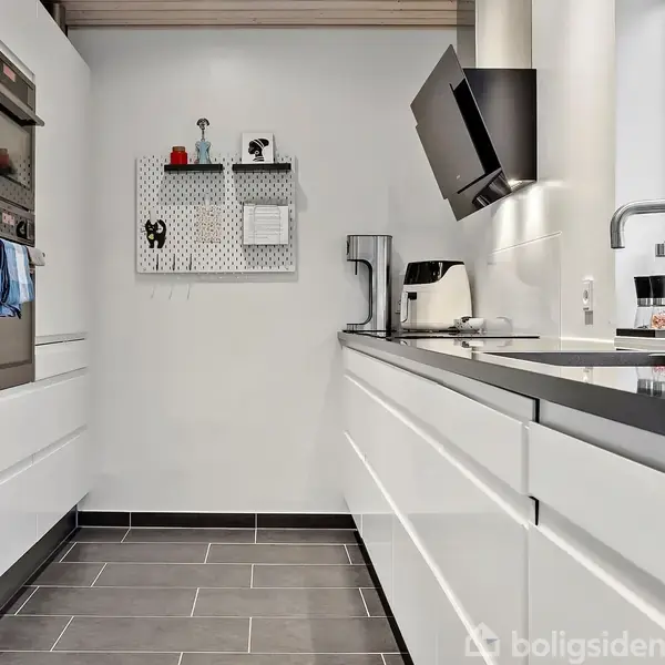 Et moderne køkken med hvide skabe og skuffer på begge sider. Et vægmonteret tv hænger over en kaffemaskine. Store vinduer giver udsigt til tage og blå himmel.