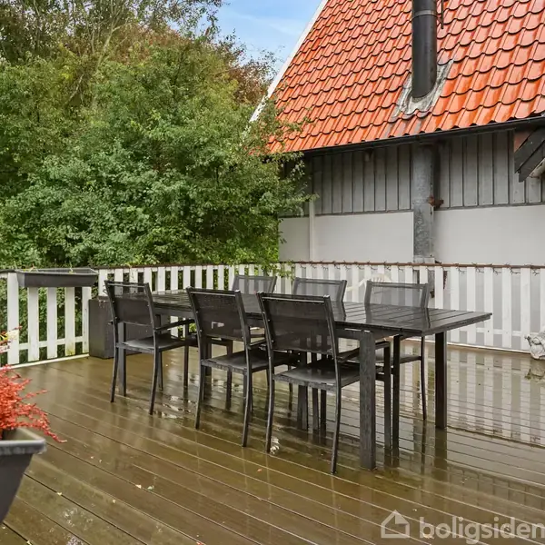Terrassemøbler står på en træterrasse med et moderne hegn. En grill er placeret i nærheden. Omkring er der frodig vegetation og et hus med rødt tegltag.