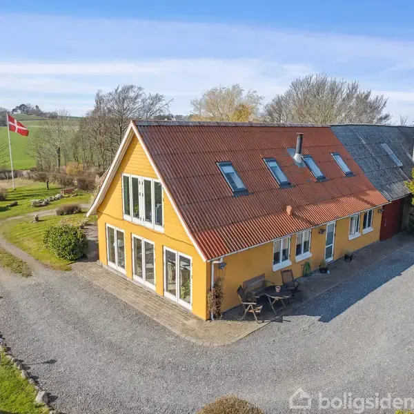 Et gult hus med rødt tag har store vinduer og en terrasse med borde og stole. Det står på en grusvej omgivet af grønne marker og en flagstang med Dannebrog.