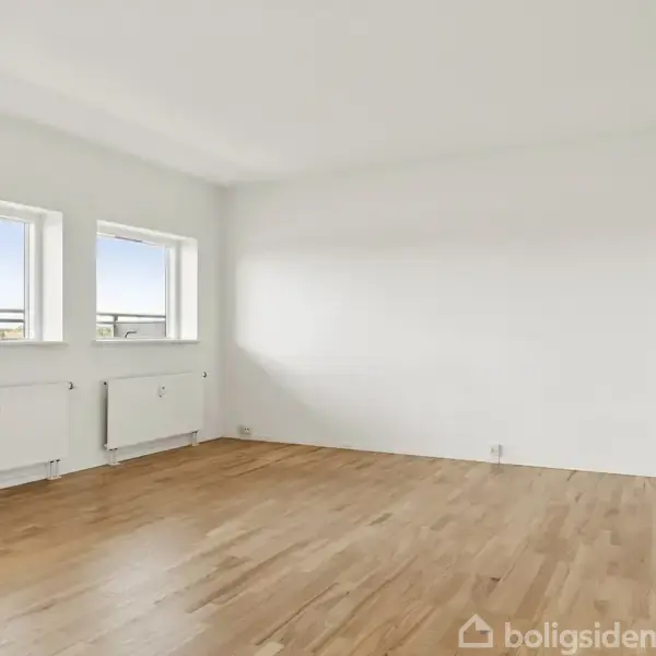 Et tomt rum med trægulv, hvor sollys strømmer ind gennem to vinduer på væggen til venstre. Der er en radiator under hvert vindue.