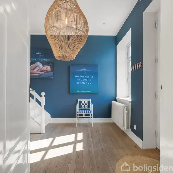En stor hvid lampe hænger fra loftet i en gang med trægulv og blå vægge. En stol står ved væggen ved siden af teksten "PUT THE HUSTLE AND BUSTLE BEHIND YOU".