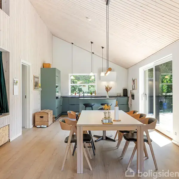 Et lyst køkken-alrum med spisebord og seks stole. Bordet er dekoreret med blomster. I baggrunden ses et moderne køkken, store vinduer og en lille hyggekrog til venstre med puder.