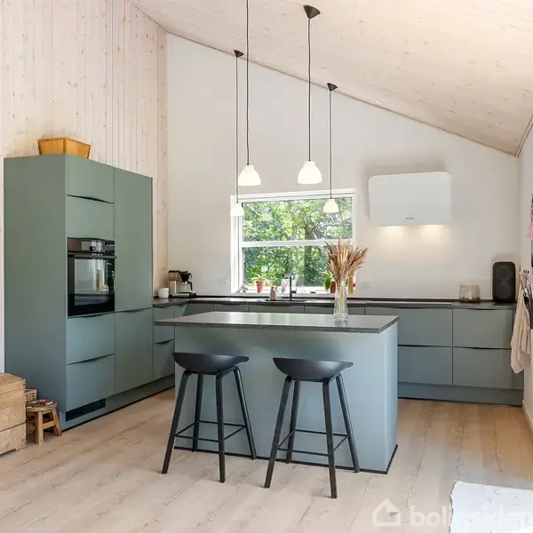 Moderne køkken med grønne skabe og ø. To høje stole står ved øen. Rummet er lyst med trævægge og gulv. Et vindue giver udsigt til træer udenfor.