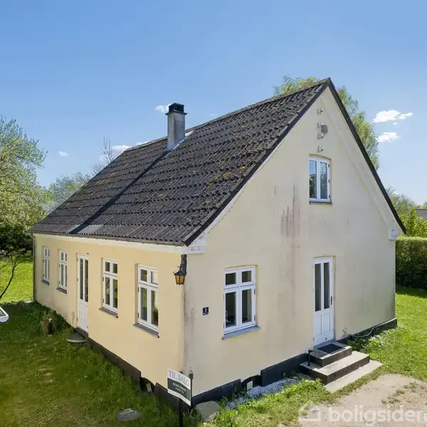 Et gult hus med sort tag står i en grøn have. Huset har hvide vinduer og en dør med trin foran. Solen skinner, og omgivelserne inkluderer træer.