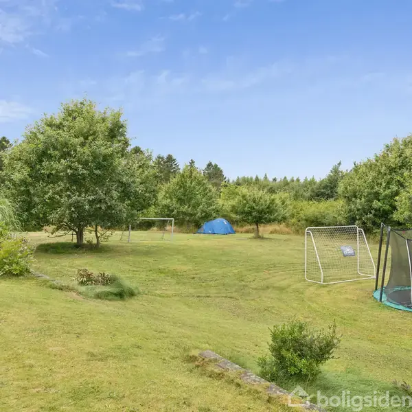 Fodboldmål og trampolin står på en stor, grøn græsplæne omgivet af træer og buske under en blå himmel.