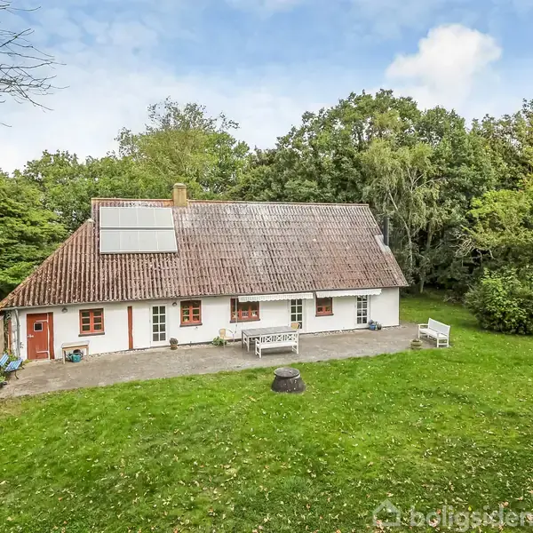 Hvidt hus med rødt tag, placeret i en grøn have med træer omkring. Huset har flere vinduer og en dør til venstre. En solcelle er monteret på taget.