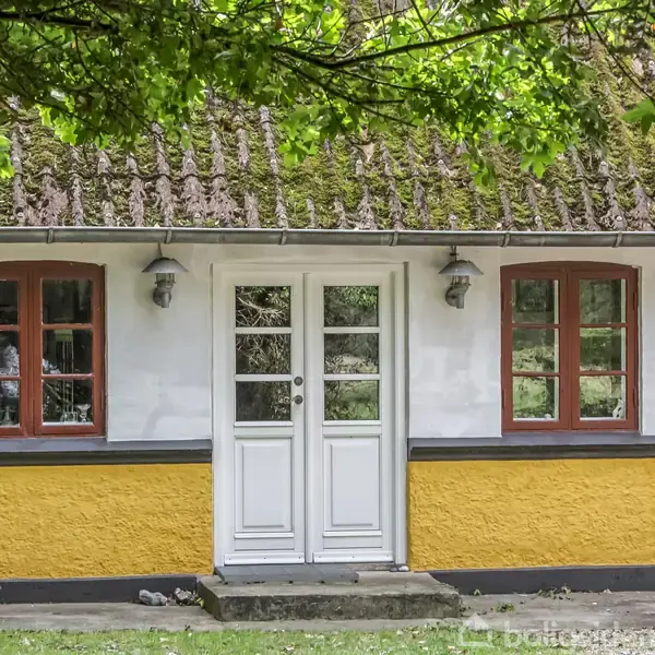 Et traditionelt bindingsværkshus med en hvid dør og rødbrune vinduer. Mosdækket tag ovenover, omkranset af grønne træer, skaber en idyllisk, landlig atmosfære.