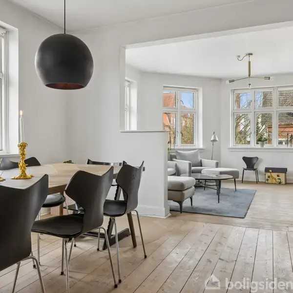 Et spisebord med sorte stole står i et lyst rum. Rummet har store vinduer med udsigt til bygninger udenfor og en åben forbindelse til en stue med sofa og tæppe.