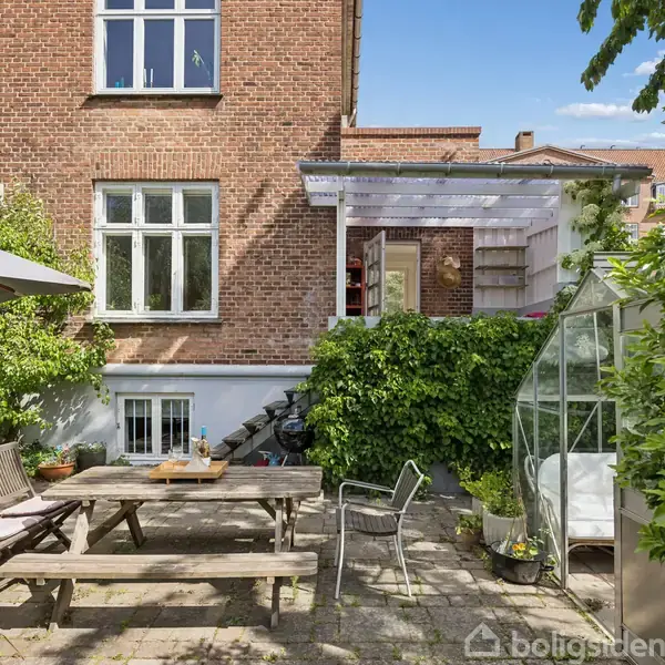 En gårdhave med træbord og stole står på en flisebelagt terrasse. En murstensbygning med hvide vinduer og en pergola omgiver det, med grønne planter og et lille drivhus ved siden af.