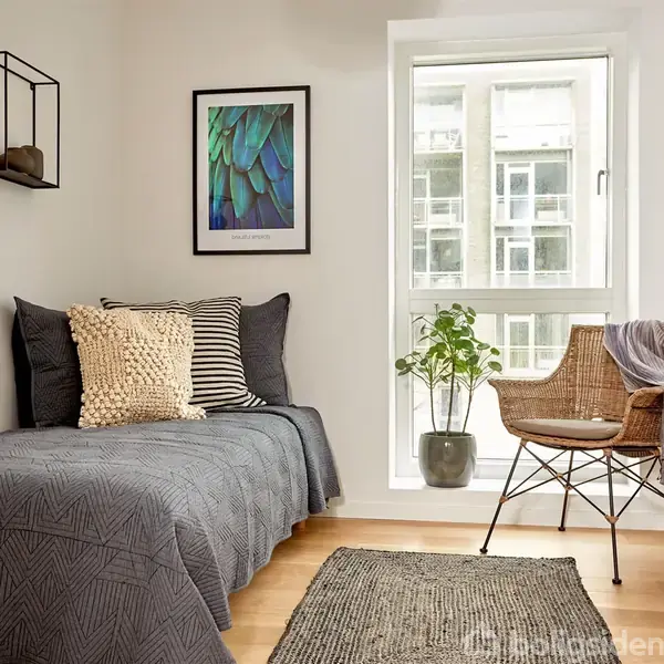 En enkeltseng dækket med gråt sengetæppe står op ad væggen under hylder med dekorationer, ved et vindue med en stol og en plante i en lys og minimalistisk stue.
