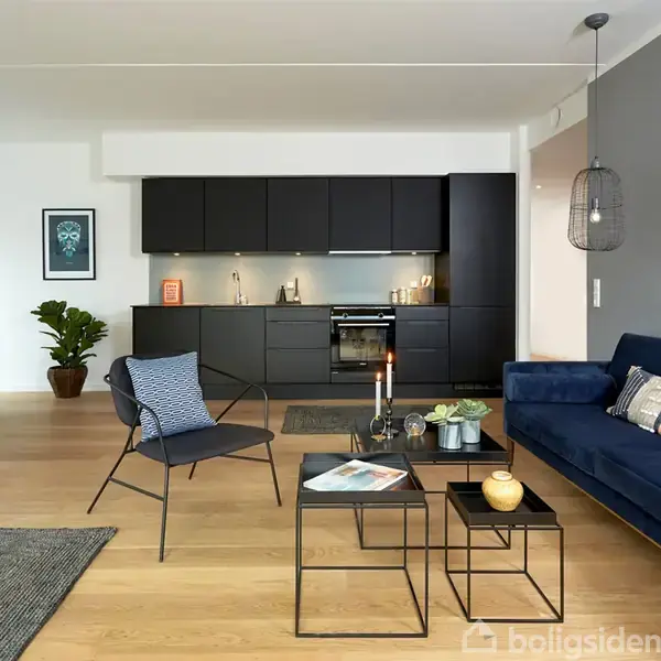 Moderne stue med blå sofa, sofaborde og stol i forgrunden; åben forbindelse til køkken med sorte skabe. Lys træparketgulve, vægkunst og planter skaber en stilfuld atmosfære.