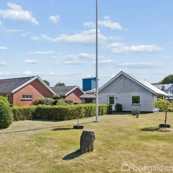 En gråmalet hus med en flagstang i forhaven står omgivet af velplejet græsplæne og buske. Flere lignende huse ses i baggrunden under en himmel med spredte skyer.