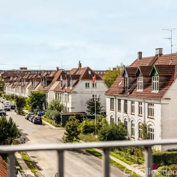 Rækkehuse med røde tage står langs en rolig, grøn gade. Biler er parkeret på vejsiden, og buske samt træer pryder fortovene i et solrigt boligkvarter.