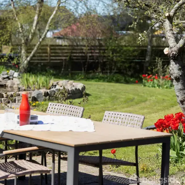 Et udendørs bord med fire flettede stole har en rød vase og en hvid dug. Det står i en frodig have med blomstrende røde tulipaner og flere træer i baggrunden.