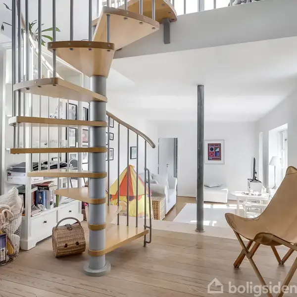 En spiraltrappe i træ og metal forbinder to etager i en lys stue med træmøbler og hvide vægge. Rummet har også en lænestol og en bogreol.